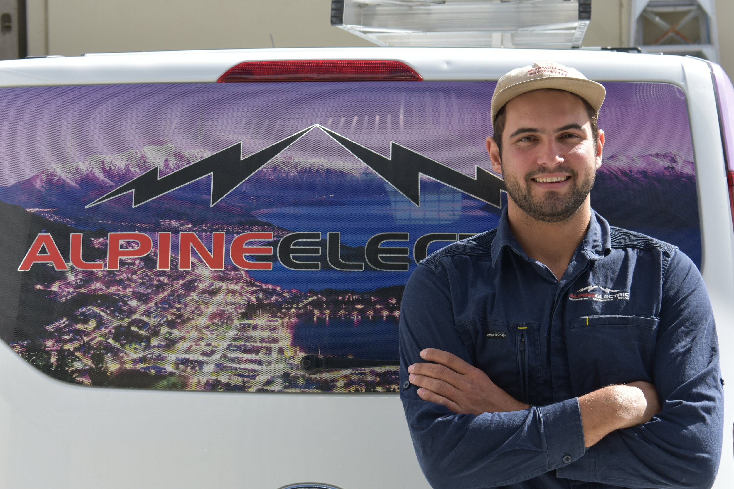 A man with crossed arms smiling in front of a van with a mountain and lake graphic, and the text 'Alpine Electric' on the back of the van.