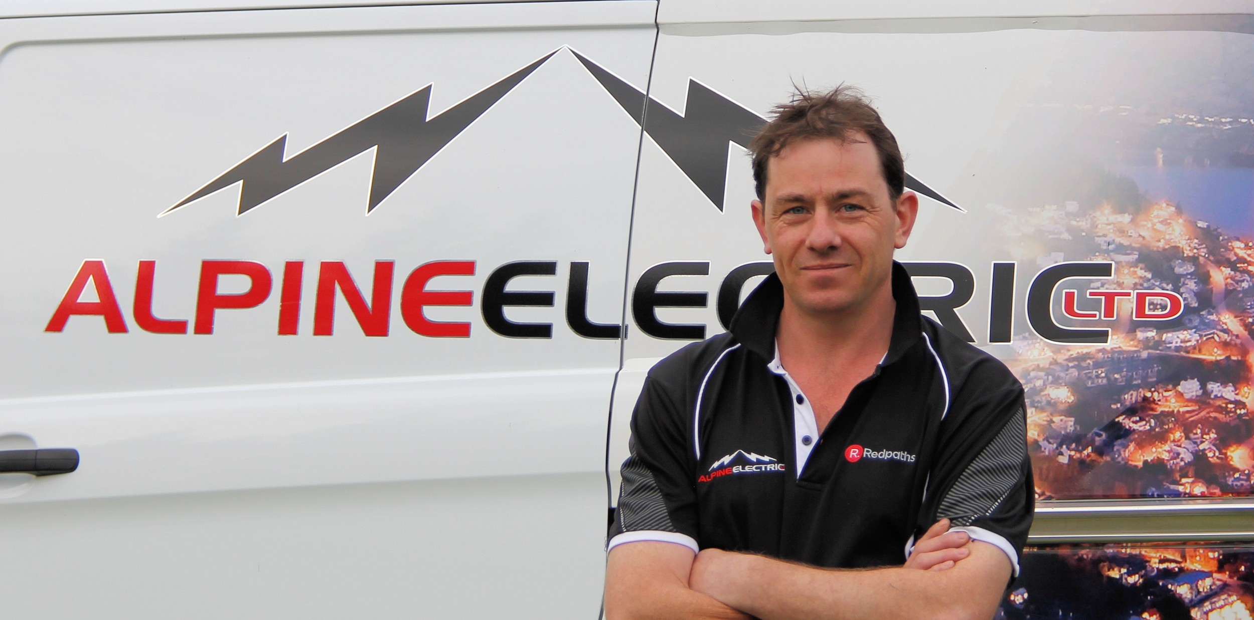 Man standing with arms crossed in front of a van with the logo 'Alpine Electric Ltd' and a mountain graphic.